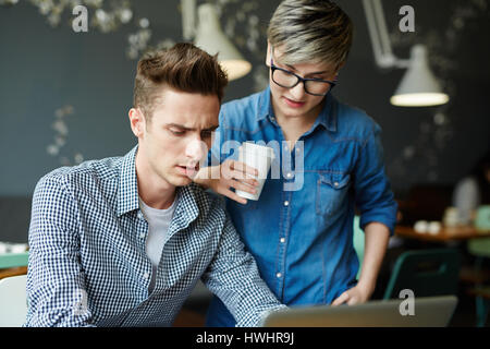 Konzentrierte junge Manager am Café-Tisch sitzen und versuchen, Probleme mit dem Computer während seine Kollegin Auge auf seine Aktionen zu lösen Stockfoto