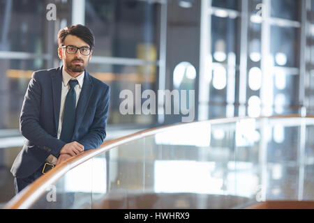 Porträt von zuversichtlich nahöstlichen Geschäftsmann mit Brille. Stehen, stützte sich auf Glas Geländer und Blick in die Kamera in der modernen Halle der Nacht Stockfoto