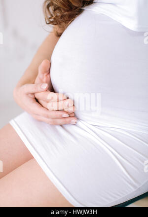 Zugeschnittenes Bild schöne schwangere Frau in einem weißen Kleid und ihr Mann umarmt den Bauch hautnah. Konzept der Schwangerschaft. Stockfoto