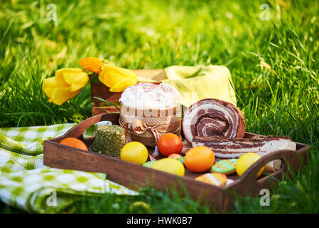 Osterkuchen und hell gefärbten Eiern gesammelt zusammen mit Fleisch, Leckereien und Tulpen auf dem Rasen unter dem Rasen Stockfoto