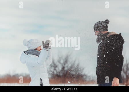 Frau in einem weißen Winterjacke, Hut und Handschuhe wirft Schnee im Angesicht ein Kerl. Sie haben eine gute Zeit auf den Winterurlaub Stockfoto