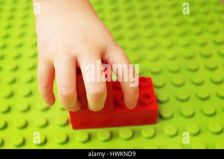 Kinder halten Konstruktor Detail auf grünem Hintergrund. Kind spielt mit Konstruktor. Kind Hand Nahaufnahme. Rote Würfel in die kleine Hand auf grünem Hintergrund. Stockfoto