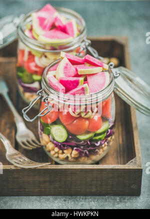 Nahaufnahme der gesunden Take-away-Gläser. Gemüse und Kichererbsen sprießen vegane Salat in Gläsern in Holztablett, grauer Beton Hintergrund, selektiven Fokus. Stockfoto