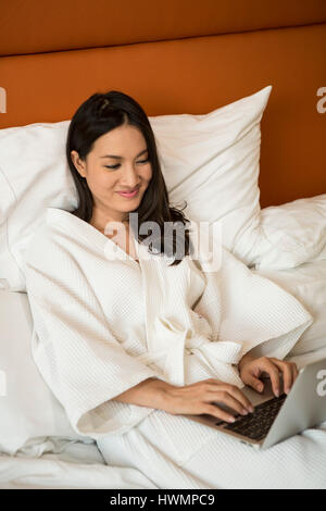 Eine asiatische Frau, die an ihrem Computer im Bett arbeitet Stockfoto