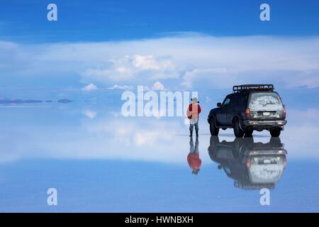 Bolivien, Potosi, Altiplano, Salar de Uyuni, Mann mit dem Hut in der Nähe seiner 4X4-Fahrzeug in die blaue Welt der Salar de Uyuni, dem größten Salzsee der Welt, mit bewölktem Himmel auf den nassen Boden wider Stockfoto