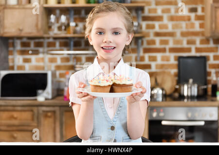 Glückliches kleines Mädchen hält köstliche Kuchen auf Teller und in die Kamera Lächeln Stockfoto