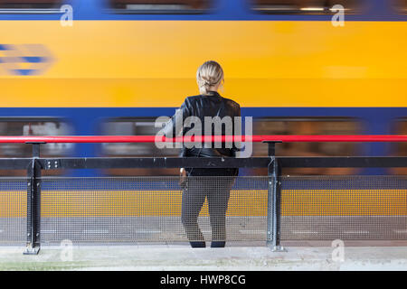 junge Frau in schwarzem Ledermantel wartet auf Plattform des Bahnhofs in den Niederlanden während gelbe und blaue Zug vorbeifährt Stockfoto