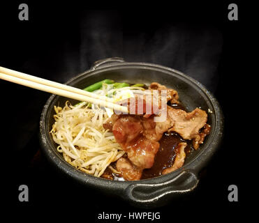 Großaufnahme Holz Shopstick halten Grill Schweinefleisch mit Sauce, Ei, Bohnen und Gemüse in heiße Pfanne asiatisch Stil in schwarzem Hintergrund schieben Stockfoto