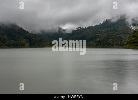 Niedrige Wolken Fahrt über den Wipfeln des Baumes bedeckt Hügel an einem großen See-Einstellung. Stockfoto