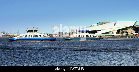 Fußgänger und Fahrrad Fähren überqueren den Fluss IJ hinter Central Station in Amsterdam, Niederlande. Auge Filmmuseum im Hintergrund Stockfoto