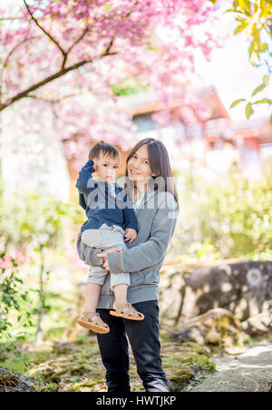 Asiatische Mutter halten Baby im Garten blühen Kirschblüten. Stockfoto