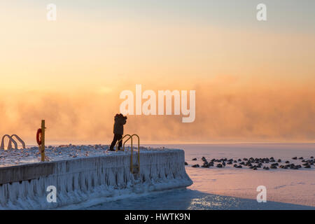 Ein Mann steht auf einem Pier bei Sonnenaufgang fotografieren Branta Canadensis (Kanadagänse) an einem sehr kalten Morgen. Oakvile, Ontario, Kanada. Stockfoto