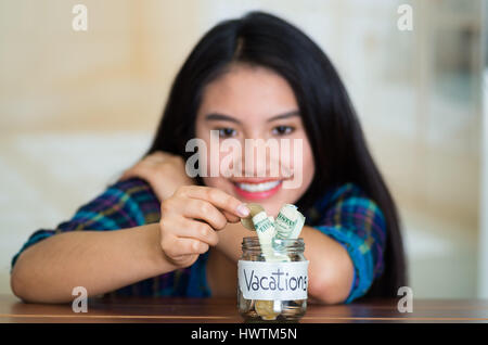 Junge Brünette Frau sitzt nach unten gerichtete Kamera, setzen Münzen in Glas mit Geld im Inneren Etikett lesen Urlaub, glücklich lächelnd Stockfoto