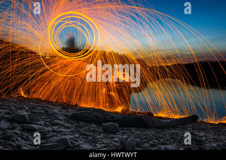 Eine Person steht auf einem Felsen gegen einen See in der Abenddämmerung Spinnerei Stahlwolle auf Feuer mit fliegen in alle Richtungen Funken. Stockfoto