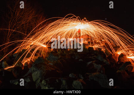 Eine Person steht hoch auf einem Hügel Spinnen Stahlwolle auf Feuer mit Funken von den Felsen in Richtung der Kamera auf den Boden Prellen. Stockfoto