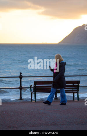 Eine einsame Frau zu Fuß entlang der beliebten Promenade in das Seebad Llandudno in Nord-Wales, wie sie zurück auf den Sonnenaufgang über den Little Orme blickt Stockfoto