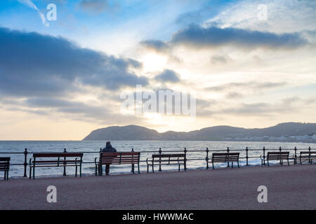 Eine Silhouette Mann sitzt auf einer Promenade Bank den Sonnenaufgang zu beobachten Pause über den Little Orme an der beliebten Küste Stadt von Llandudno in Nord-Wales Stockfoto