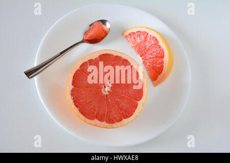 Rosa Grapefruit-Scheiben und die Hälfte auf einem weißen Teller und Hintergrund mit einem silbernen Löffel Stockfoto
