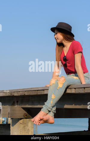 Frau trägt rote Jeans und T-shirt mit schwarzen Hut sitzt auf der Holzbrücke Stockfoto