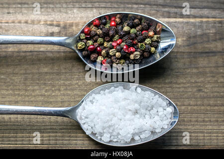 Salzkristalle und Pfefferkörner auf Metalllöffel auf hölzernen Hintergrund Stockfoto