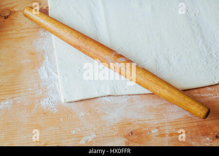 Block von frisch zubereiteten Teig auf einer bemehlten Arbeitsfläche mit einem hölzernen Nudelholz ausgerollt. Stockfoto