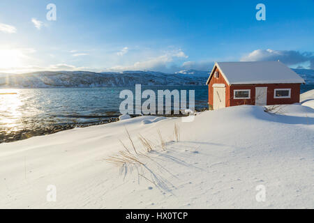 Typische norwegische warme und gemütliche Haus befindet sich am See an einem Fjord in Troms Grafschaft, Norwegen. Die Sonne ist niedrig über dem Horizont und die Landschaften eingestellt. Stockfoto