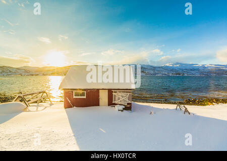 Typische norwegische warme und gemütliche Haus befindet sich am See an einem Fjord in Troms Grafschaft, Norwegen. Die Sonne ist niedrig über dem Horizont und die Landschaften eingestellt. Stockfoto