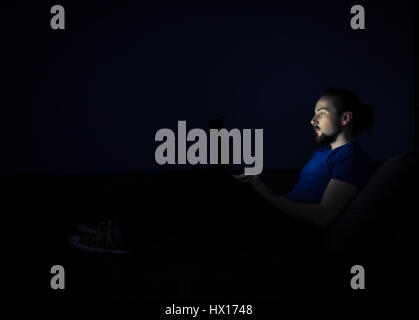 Junger Mann auf dem Bett liegend und mit Blick auf den Laptop-Bildschirm in der Dunkelheit. Stockfoto