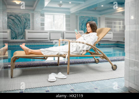 Junge Frau im Bademantel entspannen Sie sich auf die Liege in der Nähe des Beckens im Wellnessbereich im Haus Stockfoto
