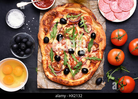 Pizza mit Tomaten, Salami und Oliven auf schwarzem Hintergrund, Ansicht von oben Stockfoto