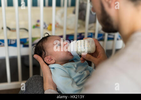 Junger Vater sitzt mit seinem Sohn in seinem Schoß. Er füttert ihn eine Flasche Milch. Stockfoto