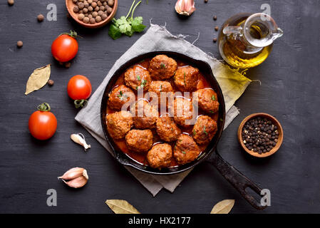 Fleischbällchen mit Tomaten-Sauce und grünen Kräutern in Pfanne auf schwarzem Hintergrund, Ansicht von oben Stockfoto