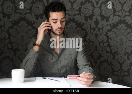 Junger Mann sitzt am Tisch zu Hause mit seinem Smartphone und seine EC-Karte, um Rechnungen über das Telefon zu bezahlen. Stockfoto