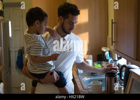 Junger Vater mache eine Tasse Tee mit Hilfe seines Sohnes. Er öffnet einen Topf mit Zucker und der kleine Knabe hält den Löffel. Stockfoto