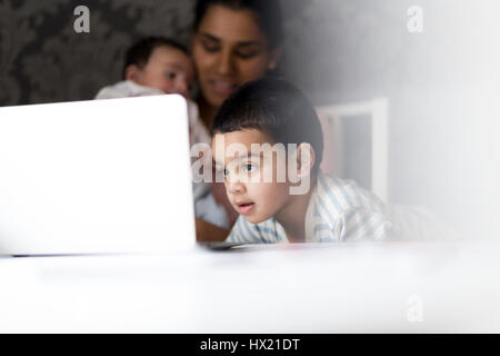 Kleiner Junge Zeichentrickfilme auf seinem Laptop Mütter während sie ihr Baby Sohn im Hintergrund füttert Stockfoto