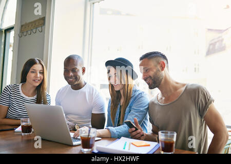 Gruppe von vier jungen Leuten mit Laptop am Schreibtisch sitzen und beobachten etwas in hellen, geräumigen Zimmer. Stockfoto