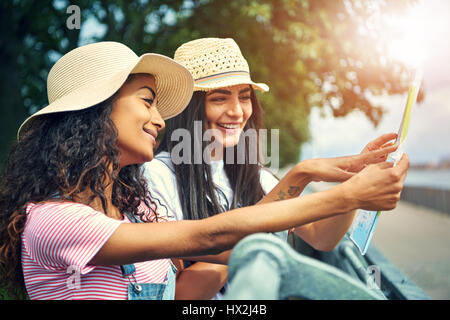 Zwei süße Freundinnen in Strohhüte lächelnd mit ihrer Karte in der hand beim schiefen gegen Zaun im freien Stockfoto