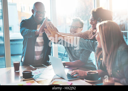 Gruppe von glücklich hohe Fiving Kollegen am Tisch im Büro. Helle große Fenster im Hintergrund. Stockfoto