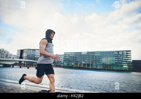 Seitenansicht des sportlichen Mann in ärmellosen Hoodie Joggen auf Asphalt der Stadt entlang des Flusses mit modernen Gebäuden im Hintergrund. Textfreiraum Stockfoto
