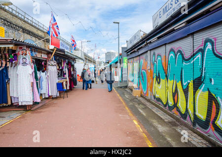 Ein Blick nach unten Shepherds Bush Market. Ein Stall auf der linken verkauft, die Kleidung während ein Geschäft auf der rechten Seite Graffiti drauf hat Fensterläden Stockfoto