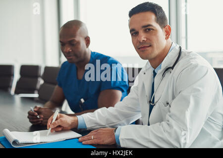 Jungen männlichen Arzt im weißen Kittel mit Stethoskop am Hals Blick in die Kamera, am Konferenztisch sitzen. Afrikanisch-amerikanischen männlichen Arzt in blau Stockfoto