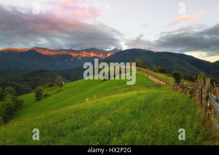 Sonnenuntergang mit grünen Wiesen und sonnenbeschienenen Gebirgszug im Hintergrund fotografiert in Siebenbürgen in der Nähe von Brasov auf einer wunderschönen hügeligen Landschaft Stockfoto