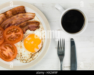 Authentische Flach bunte Englisches Frühstück, gebratenen Speck, Ei, Sunny Side Up, und gegrillten Tomaten mit einer Tasse starken Kaffee zurück Stockfoto