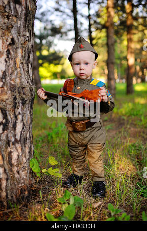 Ein nettes Kind in der Uniform eines sowjetischen Soldaten mit einem St. George Band mit einer Spielzeugpistole auf dem Hintergrund der Natur. 9. Mai, 23. Februar, Defender, aga Stockfoto