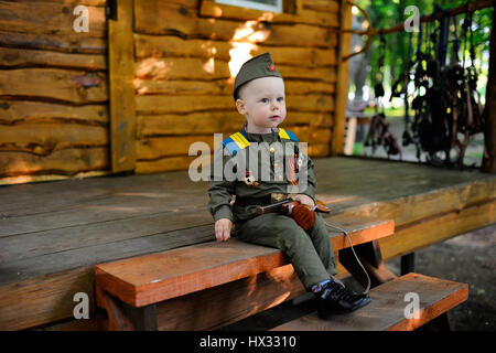 Kind in Militäruniform mit einer Spielzeugpistole auf dem Hintergrund eines Holzhauses Stockfoto
