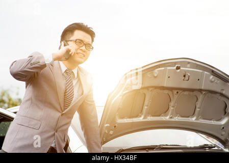 Asiatische Geschäftsleute sprechen auf einem Handy durch ein kaputtes Auto unterwegs, fordern helfen. Stockfoto