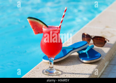 Glas Orangensaft trinken frische Blumen, Sonnenbrille und Hausschuhe am Rand eines Swimmingpools - tropischen Ferienkonzept Stockfoto