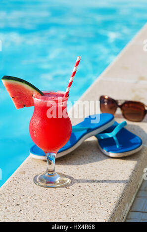 Glas Orangensaft trinken frische Blumen, Sonnenbrille und Hausschuhe am Rand eines Swimmingpools - tropischen Ferienkonzept Stockfoto