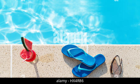 Glas Orangensaft trinken frische Blumen, Sonnenbrille und Hausschuhe am Rand eines Swimmingpools - tropischen Ferienkonzept Stockfoto