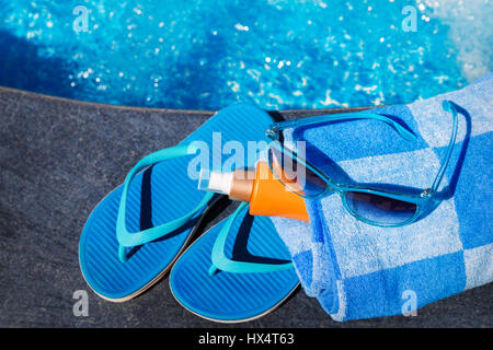 Sonnenbrille mit Sonnenschutz-Creme, blau Hausschuhe und Handtuch am Rand eines Swimmingpools - tropischen Ferienkonzept